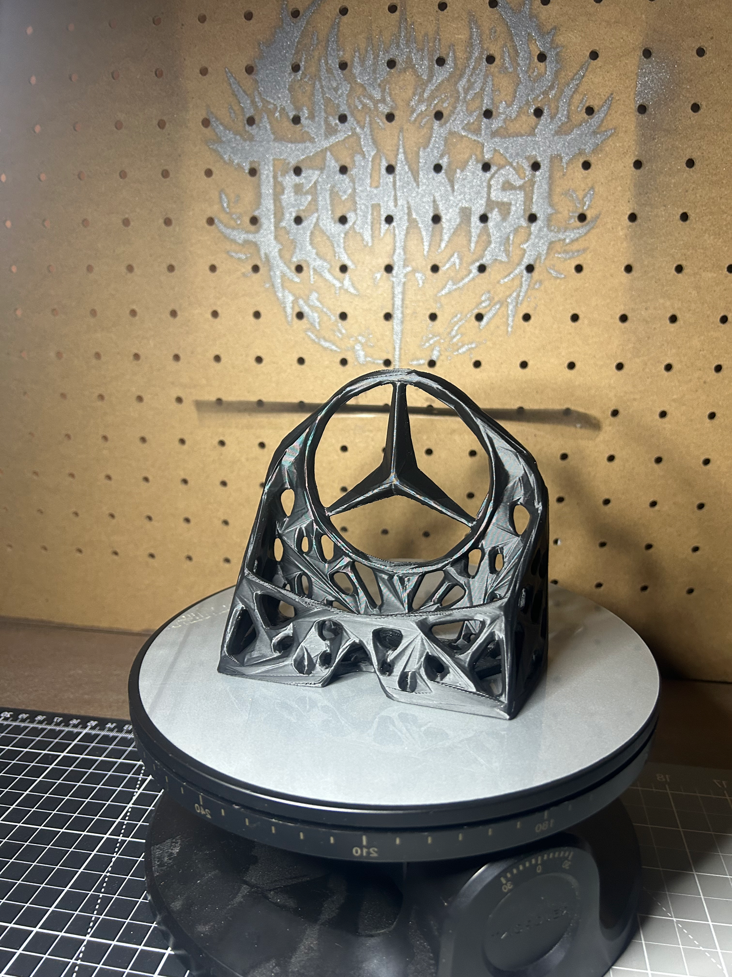 3D-printed business card stand with an avant-garde, biomorphic design featuring the Mercedes-Benz logo, displayed on a turntable in front of a pegboard with the Technast logo.