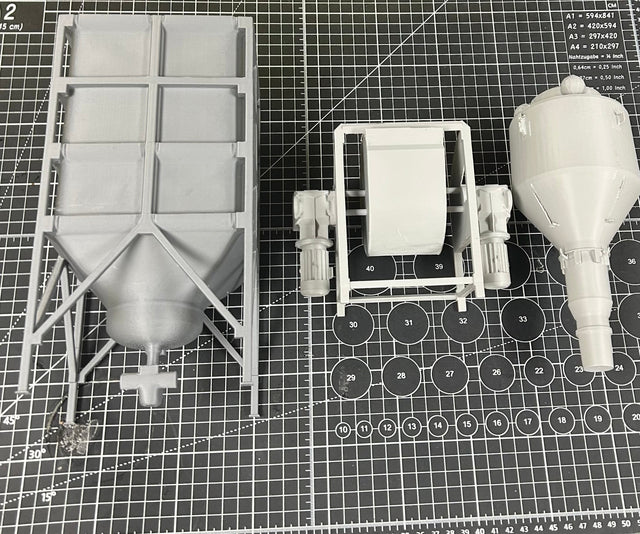 Top-down view of three large, gray 3D-printed industrial components on a measurement grid cutting mat, showcasing detail and scale. Parts include a mixer, a hopper, and a cyclone separator.